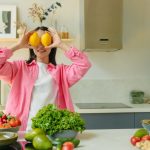 A woman playfully holding lemons over her eyes in a bright, lively kitchen setting filled with fresh vegetables.